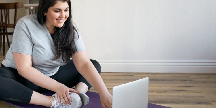 Eine übergewichtige Frau macht Fitness-Übungen vor einem Laptop.