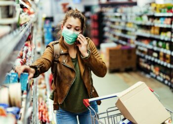 Frau mit Mund-Nasen-Bedeckung telefoniert während des Einkaufens im Supermarkt