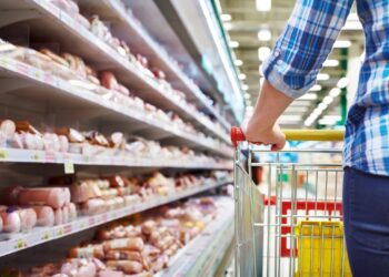 Eine Person schiebt einen Einkaufswagen an einem Wurstregal im Supermarkt entlang.