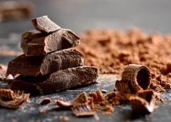 Schokoladenstücke, Schokopulver und Schokoraspeln liegen auf einer dunklen Oberfläche.
