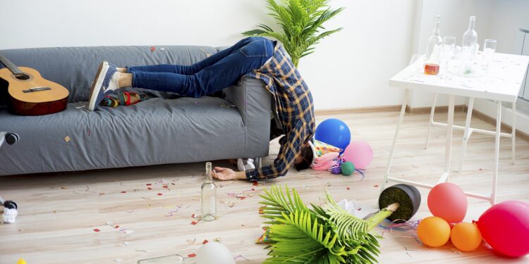 Ein Mann schläft halb aus dem Sofa hängend in einem unordentlichen Zimmer.