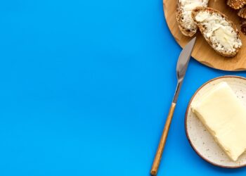 Messer, Butter und ein Brot mit Butter auf einem Holztablet vor blauem Hintergrund.
