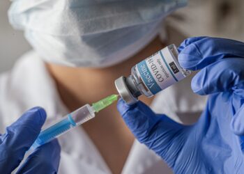 Person mit Mundschutz und blauen Handschuhen zieht einen Corona-Impfstoff in eine Spritze