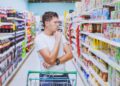 Ein Mann mit nachdenkender Körperhaltung steht vor einem Getränkeregal im Supermarkt.
