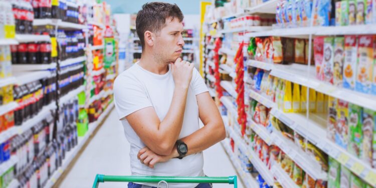 Ein Mann mit nachdenkender Körperhaltung steht vor einem Getränkeregal im Supermarkt.