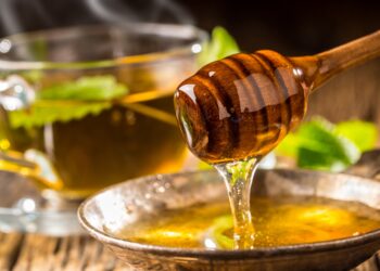 Honig und Honigschöpflöffel auf rustikalem Eichentisch und heißer Kräutertee im Hintergrund