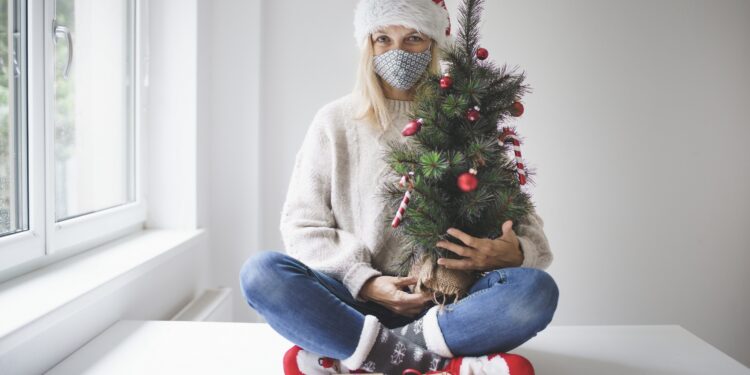 Eine Frau mit Alltagsmaske sitzt vor einem Fenster und wartet auf Weihnachten.