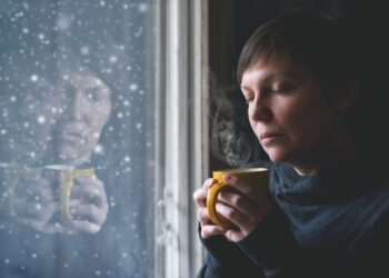 Fraurig aussehende Frau mit einer Tasse steht am Fenster