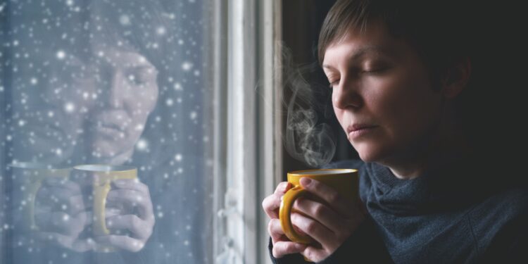 Fraurig aussehende Frau mit einer Tasse steht am Fenster