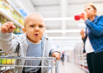 Ein Baby mit ärgerlichem Gesichtsausdruck sitzt in einem Einkaufswagen im Supermarkt.