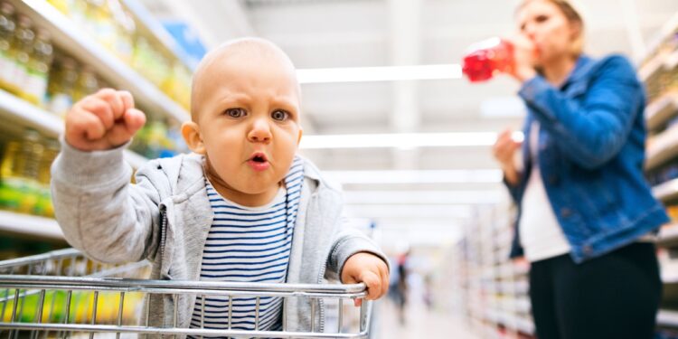 Ein Baby mit ärgerlichem Gesichtsausdruck sitzt in einem Einkaufswagen im Supermarkt.
