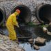 Eine Person im Schutzanzug entnimmt eine Abwasser-Probe.