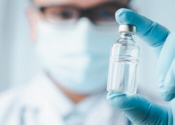 Mediziner hält Impfstoffampulle zwischen zwei Fingern