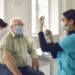 Älterer Mann mit Mund-Nasen-Bedeckung wartet auf eine Impfung, während eine Ärztin die Spritze vorbereitet