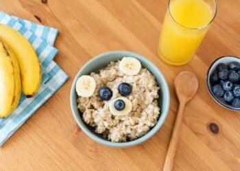 Eine Schüssel mit Porridge und Früchten sowie ein Glas Orangensaft, ein Kochlöffel, Blaubeeren und Bananen auf einem Tisch