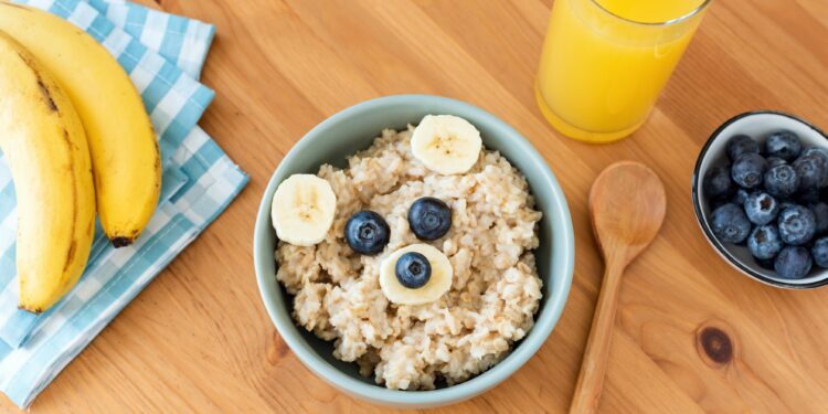 Eine Schüssel mit Porridge und Früchten sowie ein Glas Orangensaft, ein Kochlöffel, Blaubeeren und Bananen auf einem Tisch