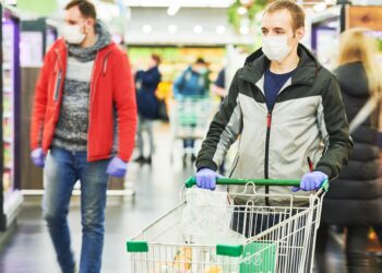 Mann mit Mund-Nasen-Bedeckung und Handschuhen mit Einkaufswagen im Supermarkt