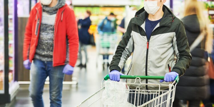 Mann mit Mund-Nasen-Bedeckung und Handschuhen mit Einkaufswagen im Supermarkt