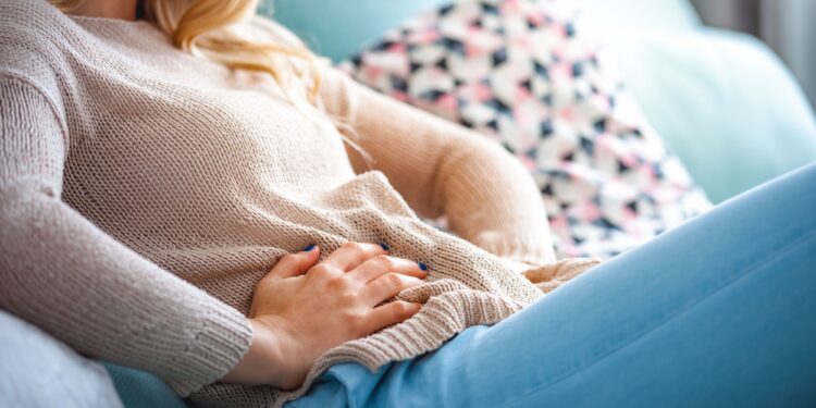 Eine junge Frau sitzt auf dem Sofa und hält ihre Hände an ihren schmerzenden Unterleib