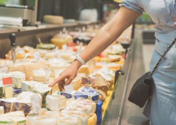 Eine Frau steht vor einer Käsetheke und zeigt auf einen Käse in der Auslage.