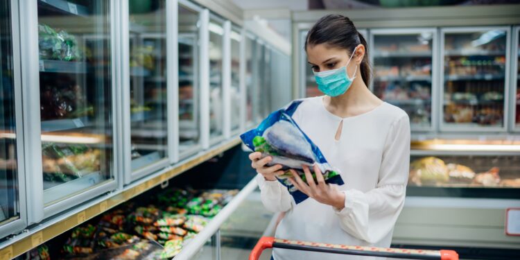 Frau mit OP-Maske liest Beschreibung eines Tiefkühlprodukts im Supermarkt