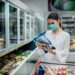 Frau mit OP-Maske liest Beschreibung eines Tiefkühlprodukts im Supermarkt