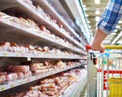 Eine Person schiebt einen Einkaufswagen entlang eines Wurstregals im Supermarkt.