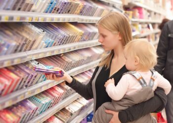 Eine Frau mit einem Baby auf dem Arm nimmt eine Schokolade aus einem Regal im Supermarkt.