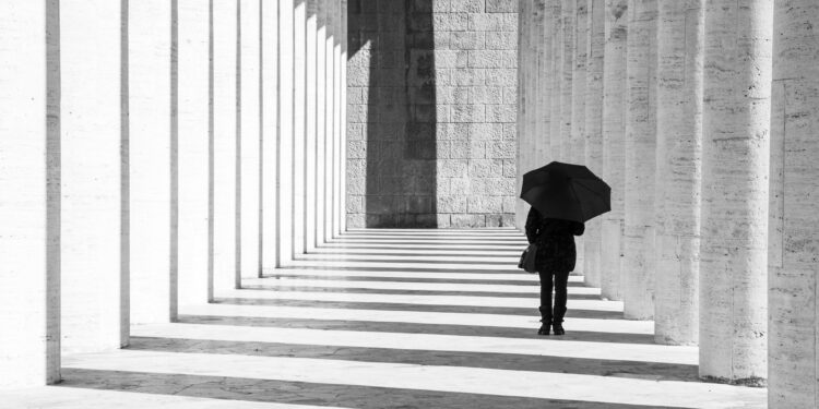 Mann in Schwarz gekleidet mit schwarzem Regenschirm geht einen Gang lang.