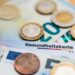 Eine Krankenkassenkarte liegt zwischen einigen Geldscheinen und Münzen.