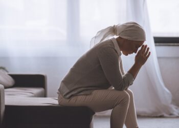 Eine Frau mit einem Tuch auf dem Kopf sitzt in niedergeschlagener Körperhaltung auf dem Sofa.