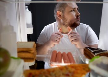 Ein Mann steht vor einem Kühlschrank und isst ein Würstchen.