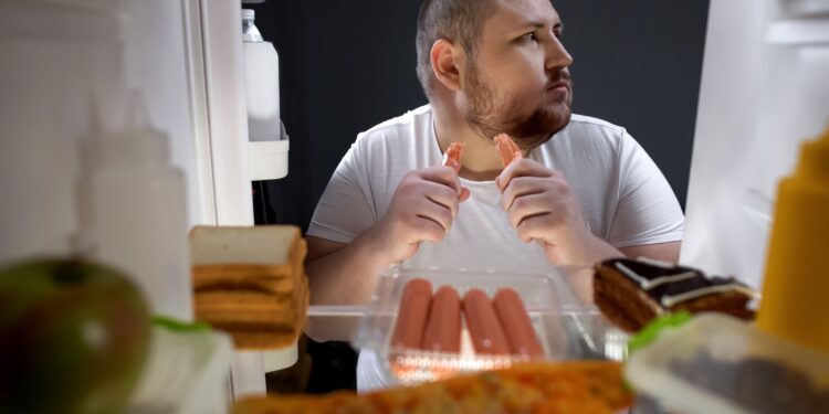 Ein Mann steht vor einem Kühlschrank und isst ein Würstchen.