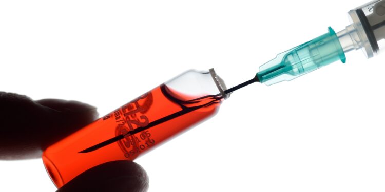 Eine Spritze zieht rote Flüssigkeit aus einem kleinen Glasfläschchen.