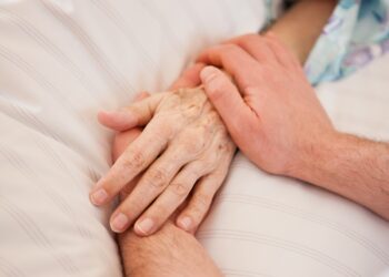 Die Hände einer Person halten die Hand einer im Bett liegenden Person.