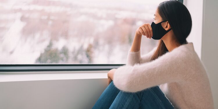 Eine Frau mit Mundschutz sitz vor einem Fenster und schaut nach draußen.