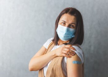 Frau mit medizinischer Maske zeigt ihr Pflaster auf dem Oberarm