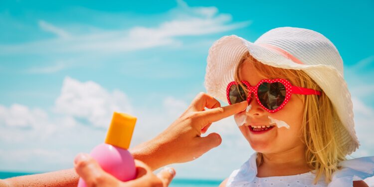 Frau cremt Mädchen mit Sonnenhut und Sonnenbrille das Gesicht mit Sonnenschutzcreme ein.