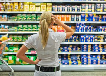 Eine Frau steht mit nachdenklicher Körperhaltung vor einem Regal im Supermarkt.