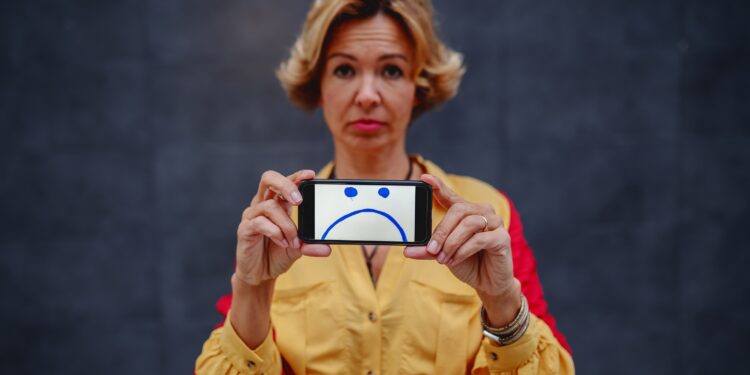 Blonde Frau zeigt Handy mit traurigem Gesicht.