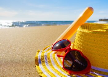 Ein Hut, eine Sonnenbrille und Sonnencreme liegen auf einem Strand.