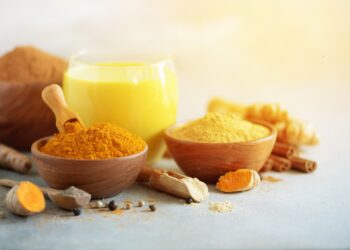 Ein Glas Goldene Milch und Holzschälchen mit Kurkumapulver, Ingwerpulver und Kurkumawurzel