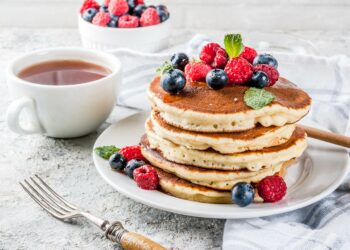 Mehrere übereinander gestapelte Pancakes sind zusammen mit Beeren auf einem weißen Teller angerichtet.