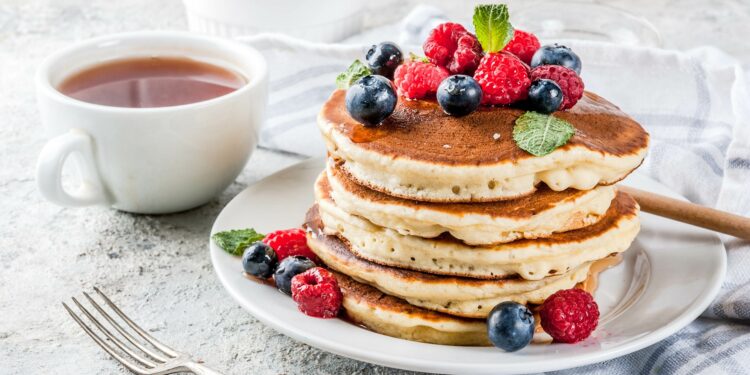 Mehrere übereinander gestapelte Pancakes sind zusammen mit Beeren auf einem weißen Teller angerichtet.