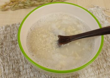Schleimige Dinkel-Hafer-Suppe in weißer Schale mit hellgrünem Rand und schwarzem Löffel auf beigefarbenem Tischset