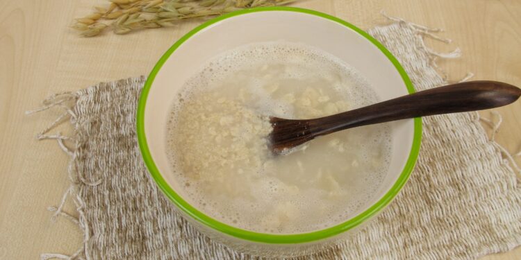 Schleimige Dinkel-Hafer-Suppe in weißer Schale mit hellgrünem Rand und schwarzem Löffel auf beigefarbenem Tischset