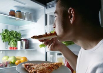 Ein junger Mann steht im Dunkeln am geöffneten Kühlschrank und isst ein Stück Pizza.