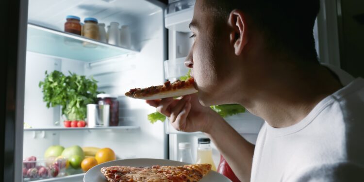 Ein junger Mann steht im Dunkeln am geöffneten Kühlschrank und isst ein Stück Pizza.