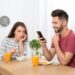 Junger Mann nutzt am Frühstückstisch sein Smartphone während ihn seine Partnerin unzufrieden ansieht