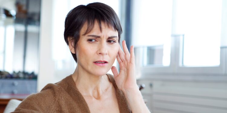 Eine Frau mit besorgtem Gesichtsausdruck fasst sich mit der Hand ans Ohr.